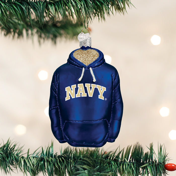 A Christmas tree ornament shaped like a navy blue hoodie with NAVY written on the front, hanging on a tree branch with blurred white lights in the background.